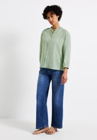 Blusa in lino verde chiaro con scollatura arricciata, abbinata a jeans in denim blu a gamba larga e sandali color cammello. Tessuto testurizzato, vestibilità casual.