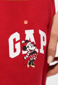 Κόκκινο ύφασμα με λευκό κείμενο GAP και τη Μίνι Μάους να φοράει καπέλο Άγιου Βασίλη, κασκόλ και φόρεμα με πουά, δίπλα στο χέρι ενός ατόμου.