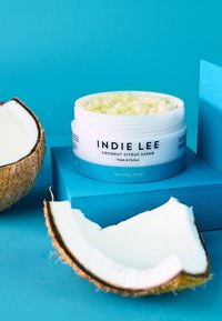 Scrub al cocco e agrumi in un barattolo bianco con etichetta blu, caratterizzato da una consistenza di scrub giallo chiaro, accanto a un cocco tagliato a metà su uno sfondo turchese.