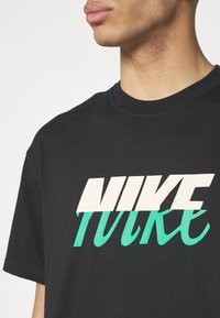 Black cotton t-shirt featuring a large, bold graphic of the word "Nike" in white and teal lettering. Crew neck and short sleeves.
