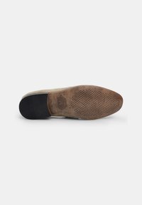 Braune Gummisohle mit strukturiertem Grip, die ein Logo-Prägung und Perforationen aufweist, kombiniert sich mit einem beigen Textil-Obermaterial für leichten Komfort.