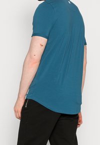 Man iförd en blå kortärmad skjorta och svarta byxor med blixtlås på sidan, sett från baksidan och sidan mot en neutral bakgrund.