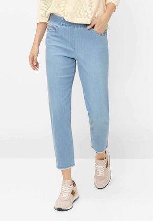 Kvinde iført lyseblå korte jeans, beige sneakers og en cremefarvet strikket sweater, stående med den ene hånd i lommen.