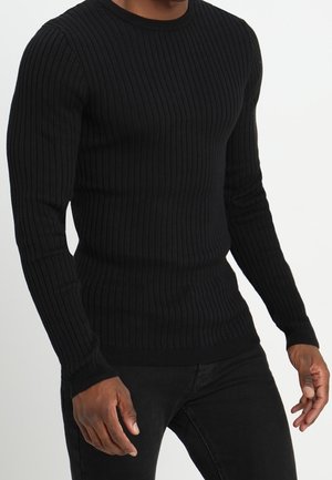 Homme portant un pull en tricot côtelé noir ajusté et un pantalon noir, se tenant contre un fond clair et uni.