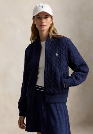 Femme portant un ensemble veste et jupe en maille torsadée bleu marine avec une chemise blanche et une casquette blanche, debout devant un fond beige uni.