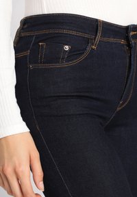 Donkerblauwe denim jeans met oranje stiksels, voorzien van een voorzakdetail en een slim fit ontwerp. Het materiaal lijkt soepel en duurzaam.