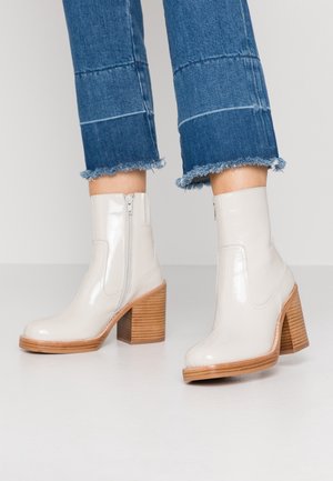 Bottes chevilles en cuir verni blanc avec un talon en bois épais, fermeture éclair sur le côté et bout arrondi. Un jean avec un ourlet effiloché complète les bottes.