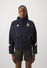 Marin zip-up hoodie med huva, vita Adidas-ränder och Real Madrid-logotyp. Matchas med olivgröna träningsshorts. Slät tygkvalitet.