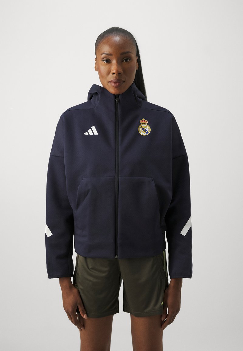 Marin zip-up hoodie med huva, vita Adidas-ränder och Real Madrid-logotyp. Matchas med olivgröna träningsshorts. Slät tygkvalitet.