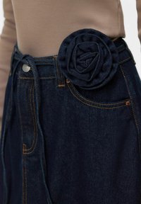 Denim seelik tumelõvilise roosikujulise kangavöö detailiga. Omab kõrge vöökohaga lõiget, ees taskut ja kontrastset oranži õmblust.