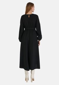 Schwarzes Langarmkleid mit tailliertem Schnitt, fließendem Rock und einem Ausschnitt auf der Rückseite. Kombiniert mit weißen kniehohen Stiefeln. Glatter Stofftextur.