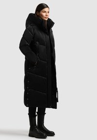 Juodas pūkuotas paltas su aukšta apykakle, su užtrauktuko uždarymu, šoninėmis kišenėmis ir mygtukais per hemą. Lygi, blizgi tekstūra.