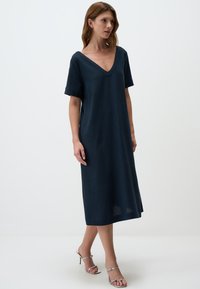 Marineblaues, knielanges Kleid mit V-Ausschnitt und kurzen Ärmeln, das eine lockere Passform und eine leinenähnliche Textur aufweist. Kombiniert mit silbernen Absatzschuhen.