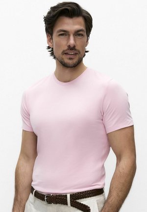 Mężczyzna o krótkich ciemnych włosach, ubrany w dopasowaną jasnoróżową koszulę z krótkim rękawem i beżowe spodnie z brązowym plecionym paskiem.