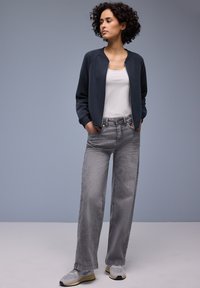 Blouson aviateur marine, débardeur blanc, jean large gris clair, et baskets grises. Textures lisses et détails matériels minimaux.