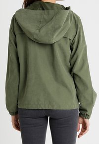 Giacca leggera di colore verde oliva, con cappuccio, polsini elasticizzati e vestibilità comoda. Texture liscia, senza motivi visibili né componenti in metallo.