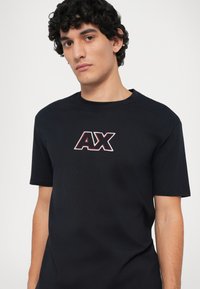 Camiseta de algodón negra con un logo "AX" en rojo y blanco en el pecho, de manga corta y diseño clásico de cuello redondo.