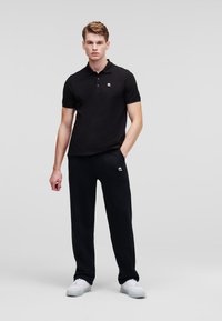 Polo nero con colletto e logo ricamato, abbinato a pantaloni neri a sigaretta e sneaker bianche su uno sfondo neutro.