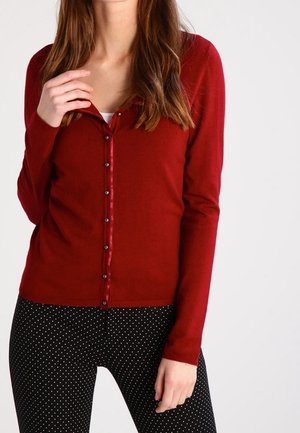 Frau trägt eine eng anliegende rote, geknöpfte Strickjacke und schwarze Hosen mit weißen Punkten, Hand nahe am Kragen, Bildausschnitt unterhalb des Kinns.