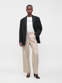 Γυναίκα που φοράει μαύρο oversized σακάκι, λευκό μπλουζάκι με γραφικά, μπεζ φαρδιά παντελόνια και καφέ ψηλοτάκουνα παπούτσια στέκεται μπροστά σε απλό φόντο.