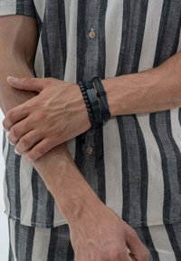 Bracelets en cuir noir et en perles portés sur une main, contrastant avec une chemise en lin grise et noire à rayures. Texturé avec des finitions lisses et mates.