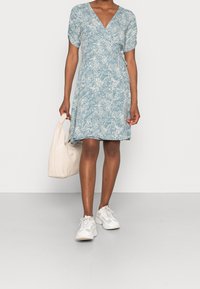 Ljusblå blommig klänning med korta puffärmar, V-ringning och utsvängd nederkant. Tillsammans med en krämliknande handväska och vita sneakers. Slät tygtextur.