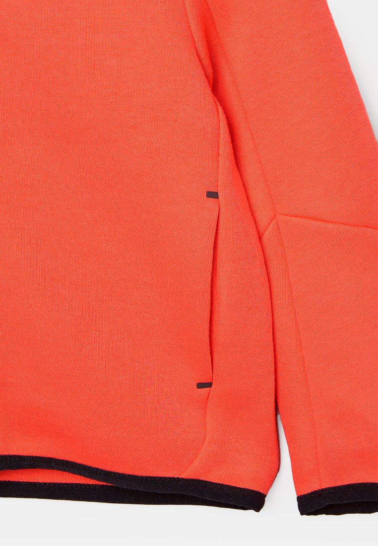 Koralowy sweter dzianinowy z jednolitą teksturą, czarnymi akcentami w szwach oraz zaokrąglonym dołem z bocznymi rozcięciami dla wygody ruchu.