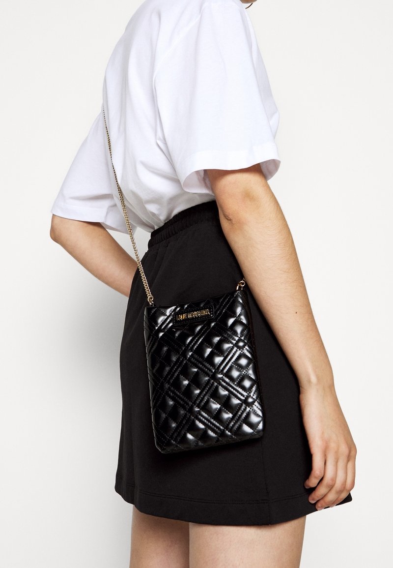 Czarna pikowana torebka na łańcuszku, z pozłacaną tabliczką z logo. Gładki, błyszczący materiał z geometrycznym wzorem.
