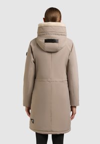 Parka imbottita beige con colletto alto, dotata di rivestimento in pelliccia sintetica, tasche laterali e dettaglio con logo sul retro, che arriva fino a metà coscia.