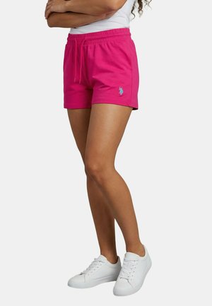 Woman wearing fuchsia drawstring shorts with an embroidered logo on the left leg, white sneakers, and a white top, standing with arms crossed.