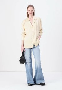 Beige skjorte med knapper og en avslappet passform, parret med lyseblå utsvingte jeans og en svart håndveske. Stoffet ser teksturert ut.