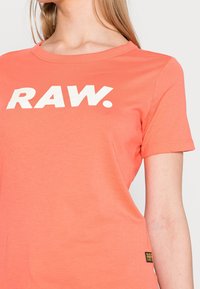 Korálová bavlněná tričko s krátkými rukávy, kulatým výstřihem a bílým grafickým nápisem "RAW." Štítek v levém dolním rohu. Hladká textura.