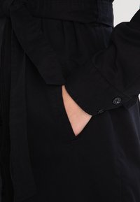 Dłoń w kieszeni czarnego płaszcza z długim rękawem, z zapięciem na guzik i materiałowym paskiem związanym w talii.
