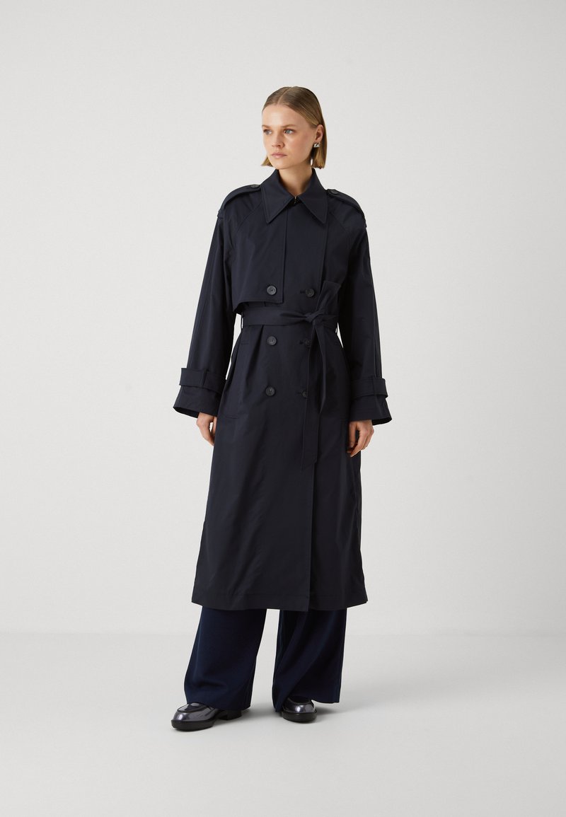 Lungo cappotto trench blu navy realizzato in tessuto liscio, con un design doppiopetto, ampio colletto e vita con cintura. Accentuato da grandi bottoni.