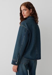 Donkerblauwe denimjas met een relaxed fit, voorzien van een klassieke kraag, knoopmanchetten en zichtbare stikdetails.