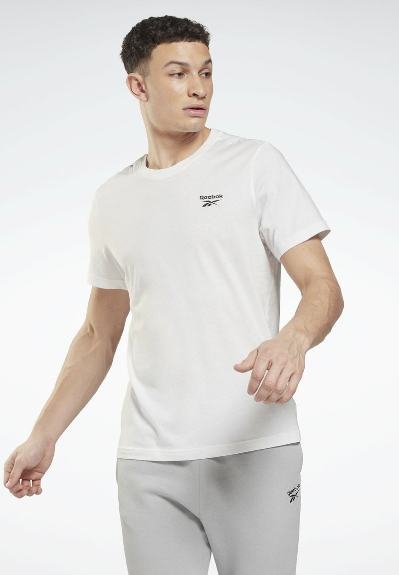 Balta kokvilnas T-krekls ar mazu melnu Reebok logotipu uz krūtīm, kombinēts ar gaiši pelēkām treniņbiksēm, izstādīts uz vienkārša fona.