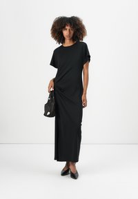 Black maxi dress with short sleeves, featuring a twisted detail at the waist. Paired with a small black handbag and pointed flats.