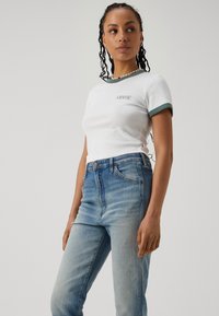 Bijela majica kratkih rukava s zelenim obrubom i Levi's logom, uparena s plavim trapericama visokog struka koje imaju izblijedjeli izgled i prednje džepove.