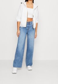 Witte lichte capuchonjas met zwarte hardware, gecombineerd met lichtblauwe jeans met hoge taille en wijde pijpen, en witte sneakers.