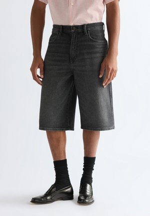 Mann trägt schwarze Jeans-Longshorts, schwarze Socken und schwarze Leder-Loafers, steht vor einem schlichten weißen Hintergrund.