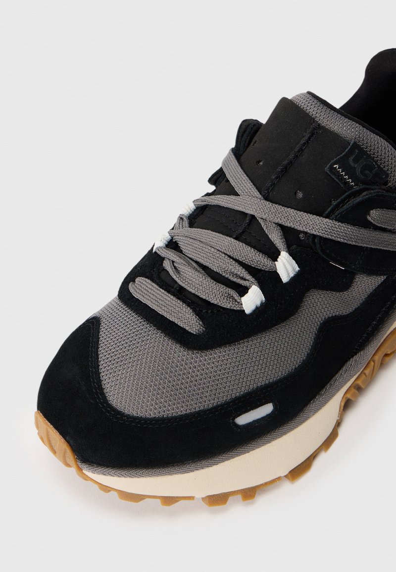 Sneaker noir et gris mettant en avant un mélange de mesh et de daim, avec des lacets gris, une semelle épaisse et une semelle extérieure en gomme texturée.