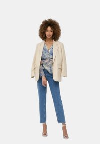 Blazer crema su una blusa blu a fantasia, abbinato a jeans dritti di lavaggio medio e tacchi a stiletto argentati. Capelli con texture, orecchini a cerchio.