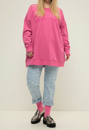 Personne portant un sweat-shirt rose surdimensionné, un jean bleu clair à motifs de cœurs roulé aux chevilles, des chaussettes roses et des mocassins compensés métalliques.