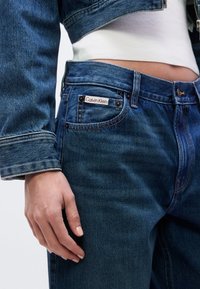 Κοντινό πλάνο ατόμου που φοράει μπλε τζιν τζιν και σακάκι με λευκό crop top, εμφανίζοντας την ετικέτα Calvin Klein στην μπροστινή τσέπη.