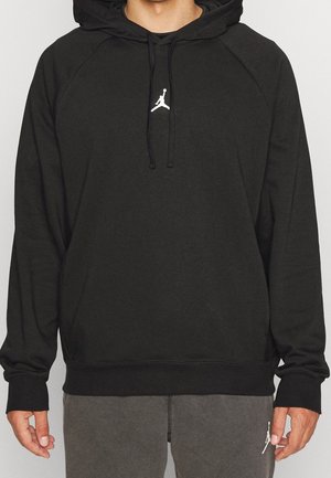 Persona che indossa una felpa nera con cappuccio con il logo Jumpman bianco sul petto e pantaloni grigi, in piedi davanti a uno sfondo semplice.