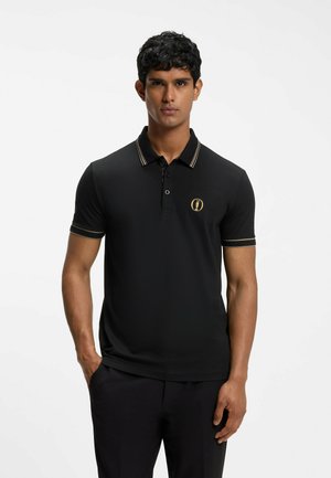 Mann steht, trägt ein schwarzes Poloshirt mit kurzen Ärmeln, goldenen Akzenten und Logo, schwarze Hose, neutraler Gesichtsausdruck, schlichter Hintergrund.