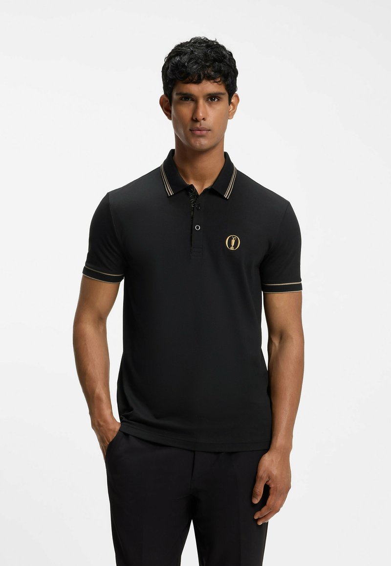 Homme debout, portant un polo noir à manches courtes avec liseré doré et logo, pantalon noir, expression neutre, fond uni.