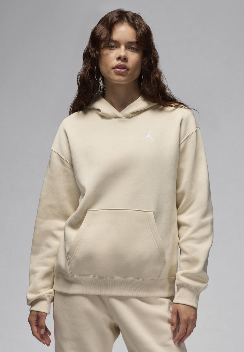 Sudadera con capucha beige, con un bolsillo frontal y puños de canalé, que presenta un pequeño logo blanco en el lado izquierdo del pecho. Textura de tela suave.