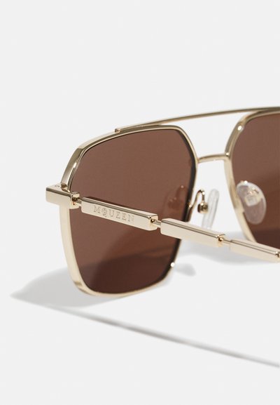 Gafas de sol con montura dorada en forma cuadrada y lentes tintados en marrón. Presenta acentos de marca y almohadillas nasales transparentes para mayor comodidad.