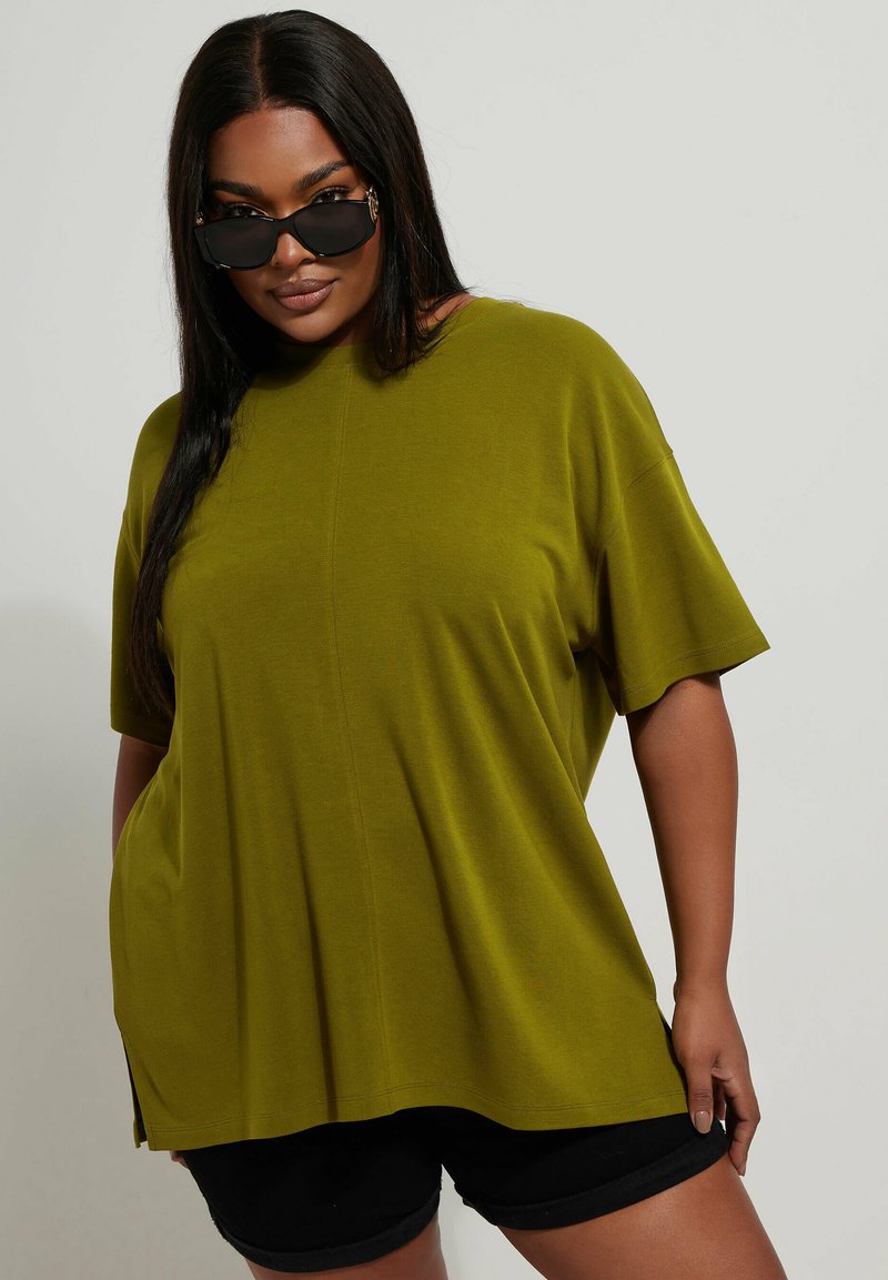 Übergroßes olivgrünes T-Shirt aus weichem Material, mit kurzen Ärmeln, rundem Ausschnitt und seitlichen Schlitzen. Tragen mit schwarzen Shorts.